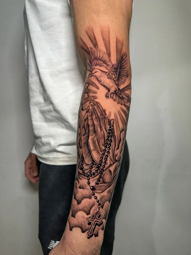 Tatuaje de manos en oración, rosario y paloma en el antebrazo
